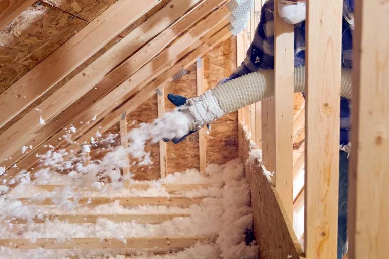 A worker spraying blown fiberglass insulation between attic trusses