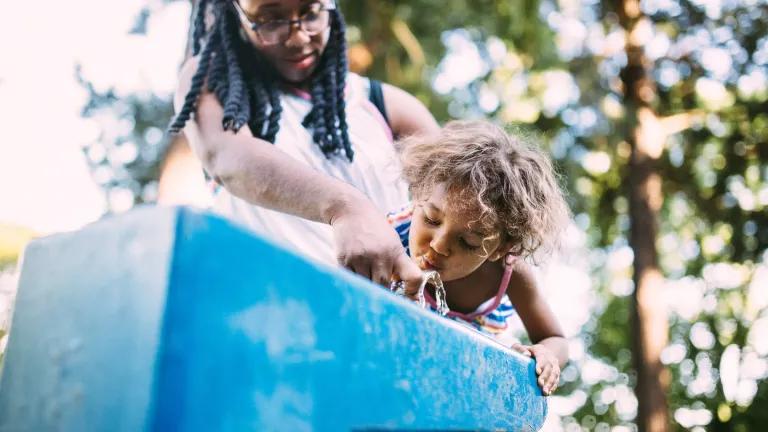 A woman lifts a young child as they drink from an outdoor water fountain