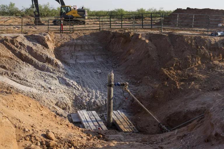 A pipe sticks out of a hole in the ground in the center of a wide pit surrounded by crude fencing
