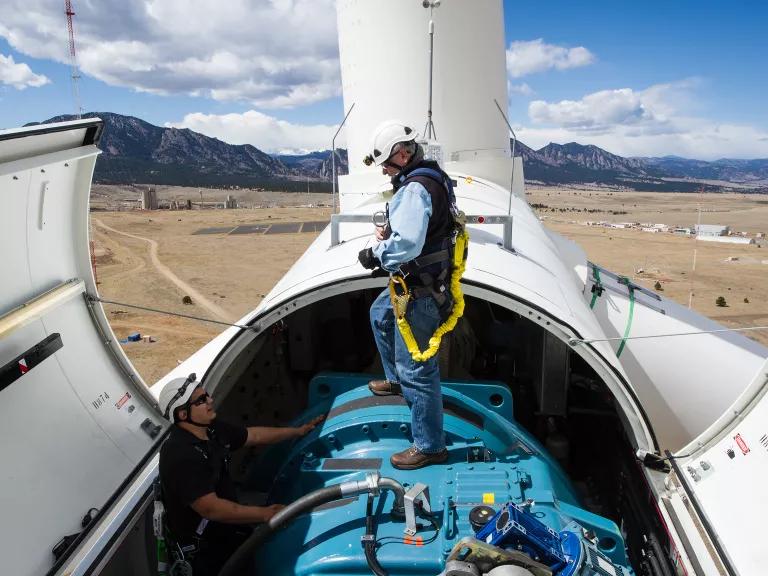 A Caucasian man in a hard hat stands on top of the nacelle of a wind turbine. The wind turbine is open, revealing the motor. Another man stands inside the nacelle, next to the motor.